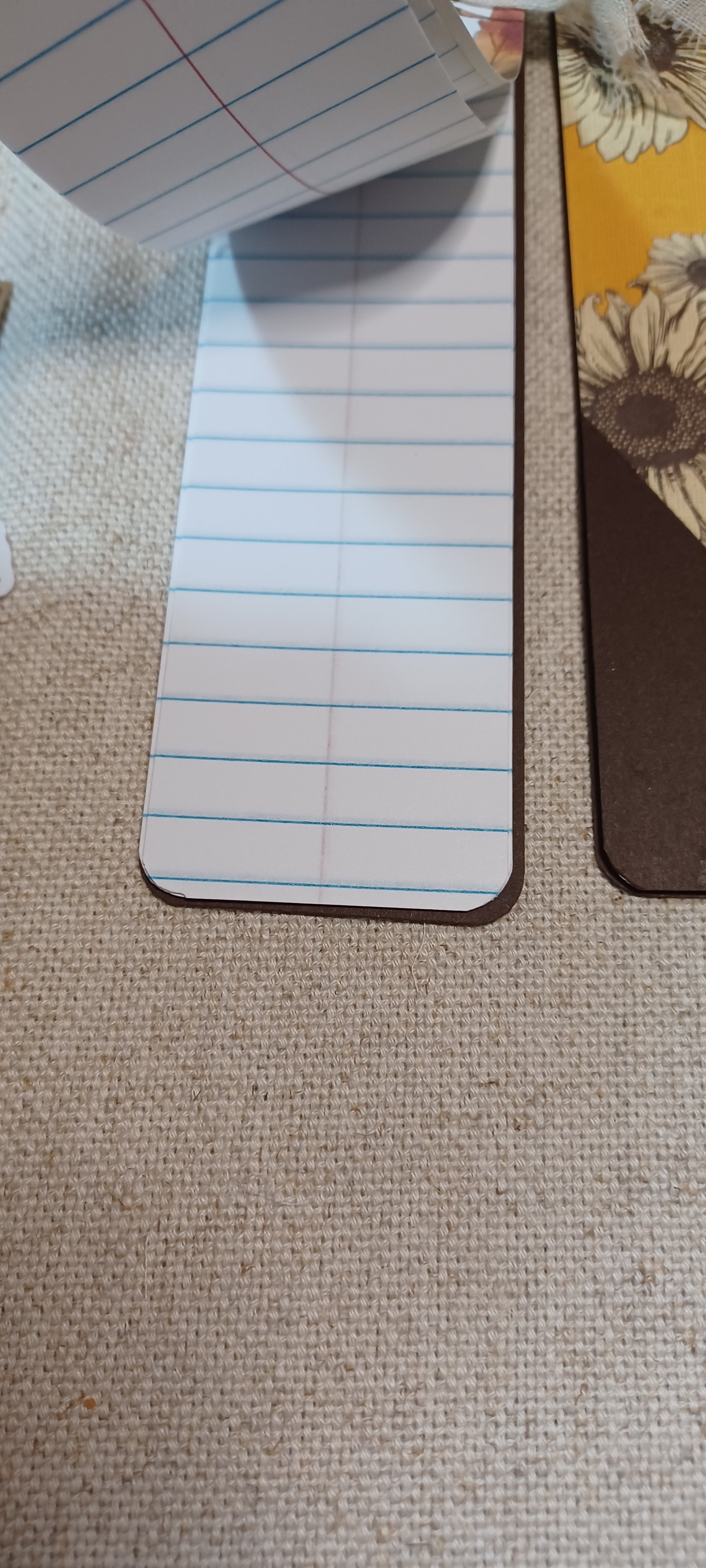 A close-up of a lined notepad sheet with blue horizontal lines and rounded corners, placed on a beige woven fabric surface next to other paper items.