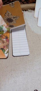A small lined notepad with a tan cover featuring a floral sunflower design, shown open on a beige fabric surface next to another floral notepad.