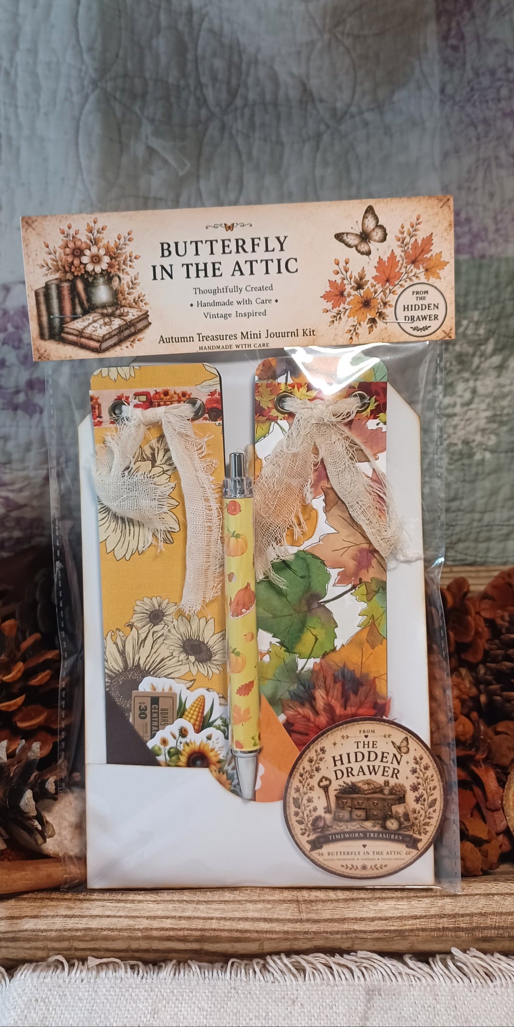 Butterfly in the Attic Autumn Transition Mini Journal Kit by The Hidden Drawer, packaged in a clear plastic bag with a decorative header card. The kit includes a journal, washi tape, and a pen, decorated with autumn-themed illustrations and ribbons.