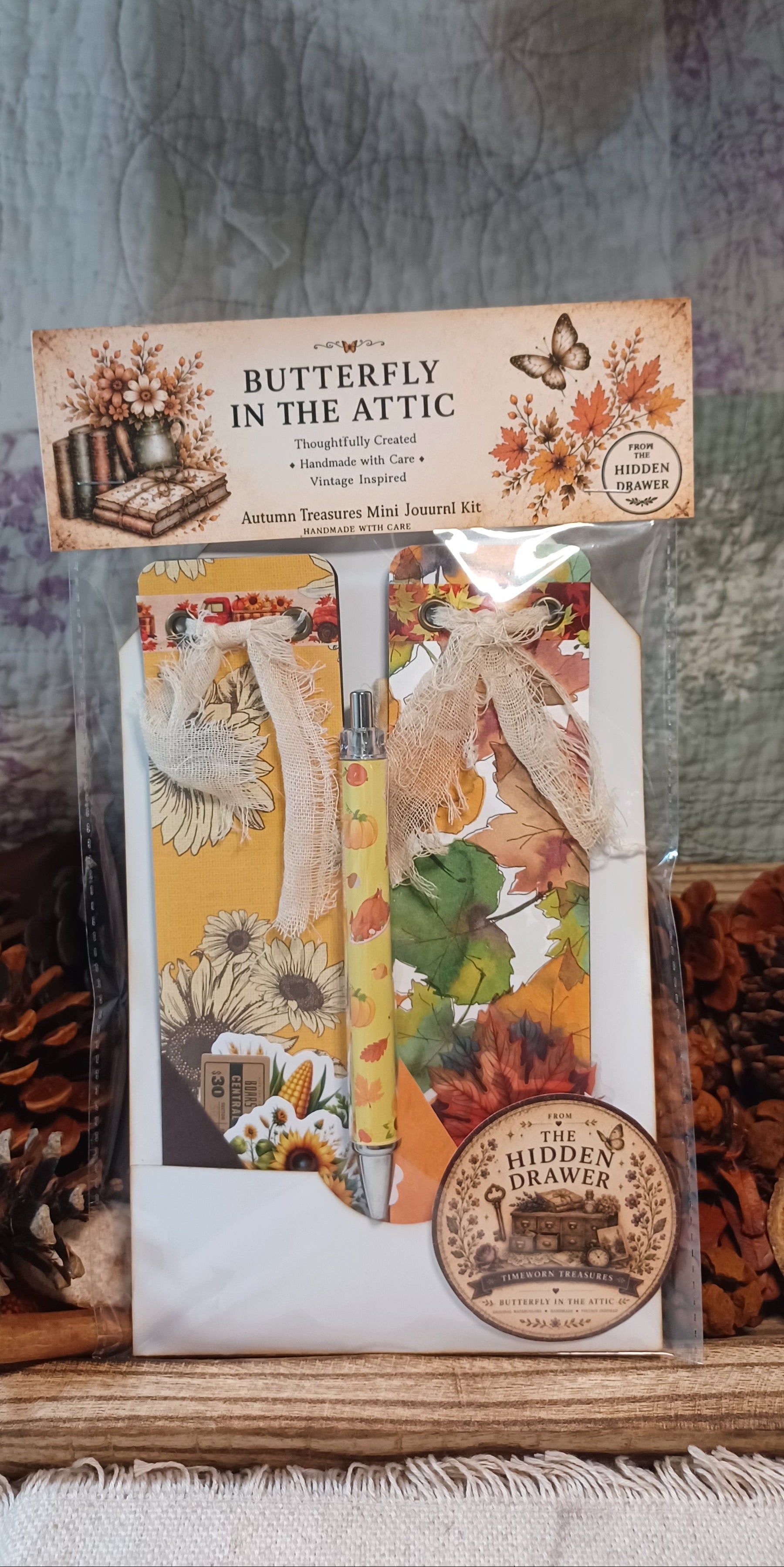 Butterfly in the Attic Autumn Transition Mini Journal Kit by The Hidden Drawer, packaged in a clear plastic bag with a decorative header card. The kit includes a journal, washi tape, and a pen, decorated with autumn-themed illustrations and ribbons.