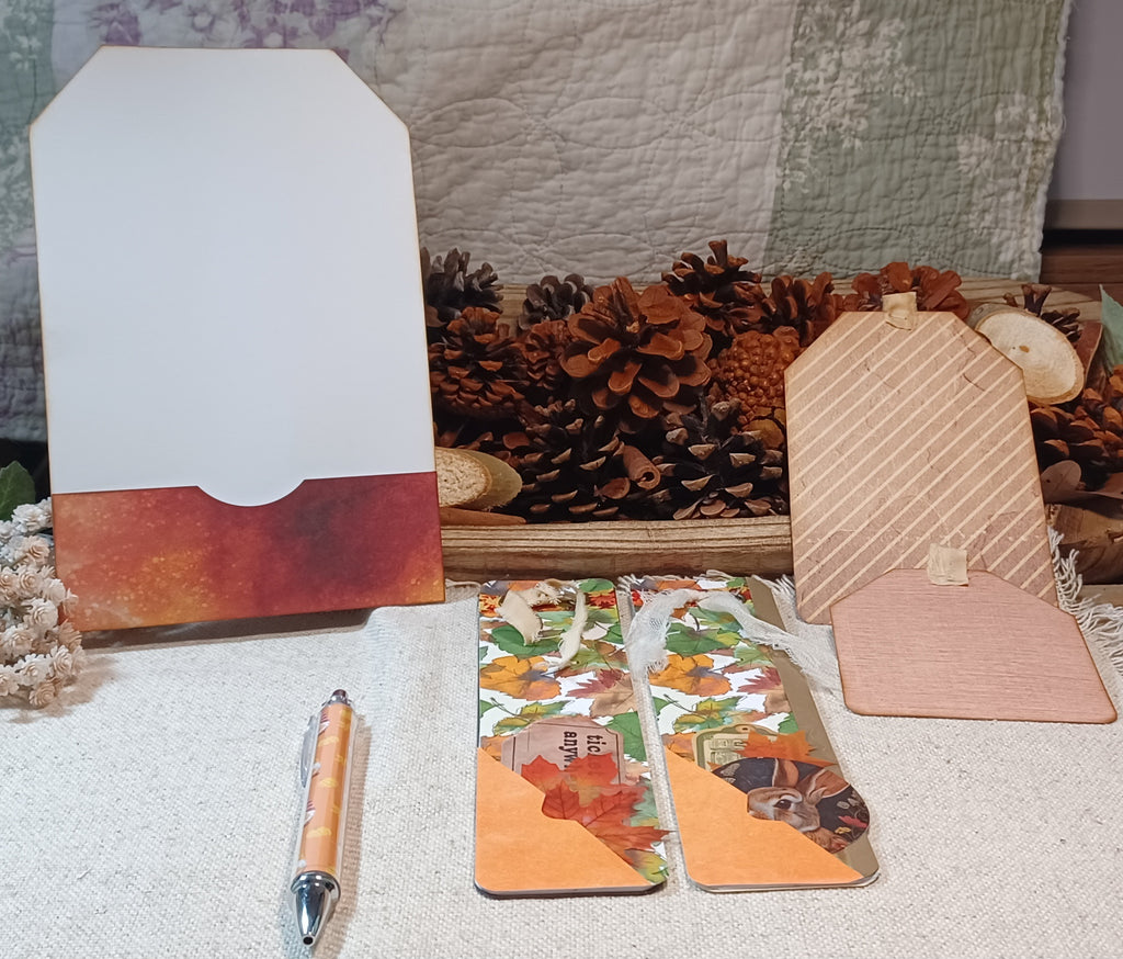 A styled flat-lay of handmade paper goods, including a white and rust-orange envelope, two patterned paper wallets with autumn leaves and pinecones, a pink paper tag, and a wooden pen, arranged on a textured white fabric with pinecones and a quilted blanket in the background.
