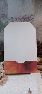 A blank white card or label with a scalloped bottom edge and a rust-orange watercolor border, displayed on a textured fabric surface with dried flowers and a rustic background.