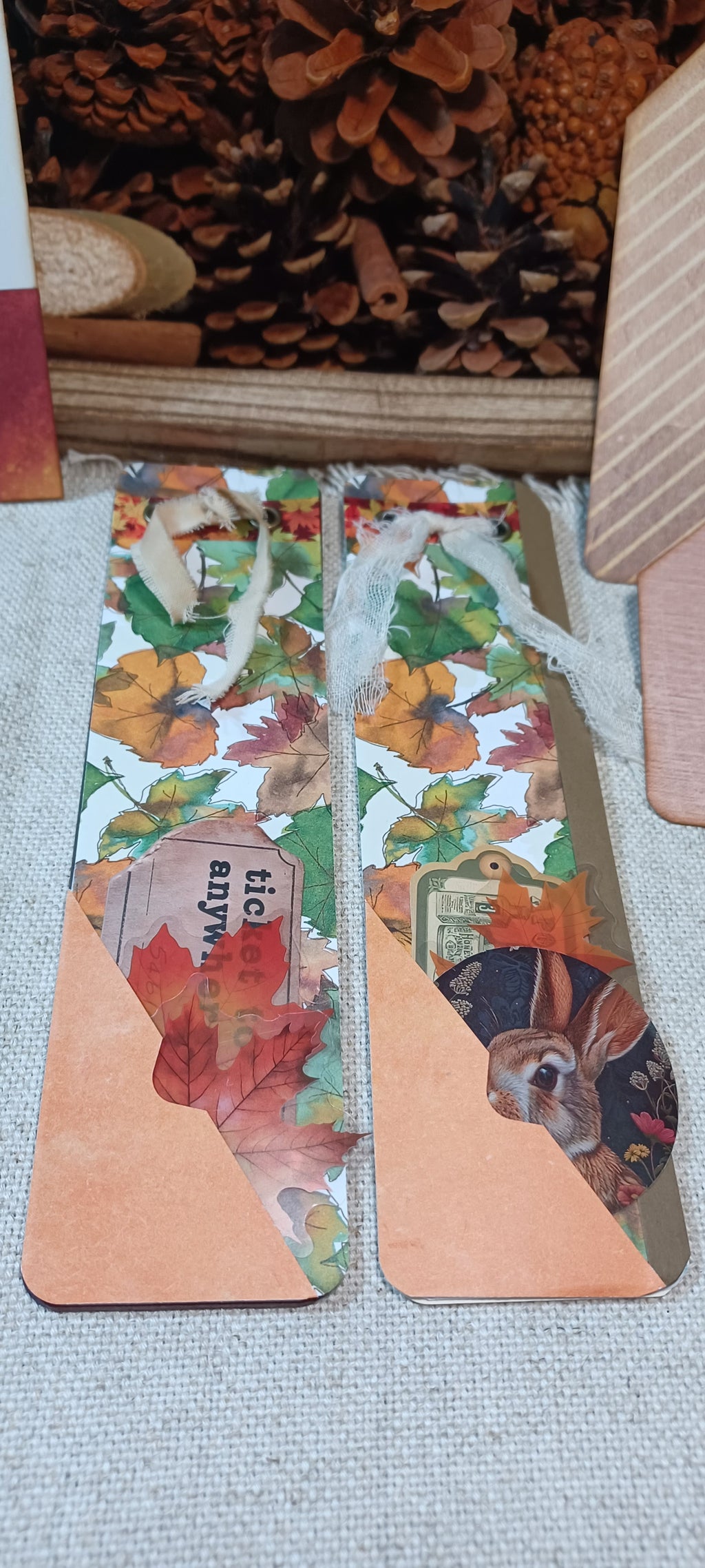 Two handmade paper bookmark holders with autumn leaf and animal collage patterns, featuring a peach-colored triangular bottom section and a white twine loop at the top, displayed on a textured gray fabric surface.