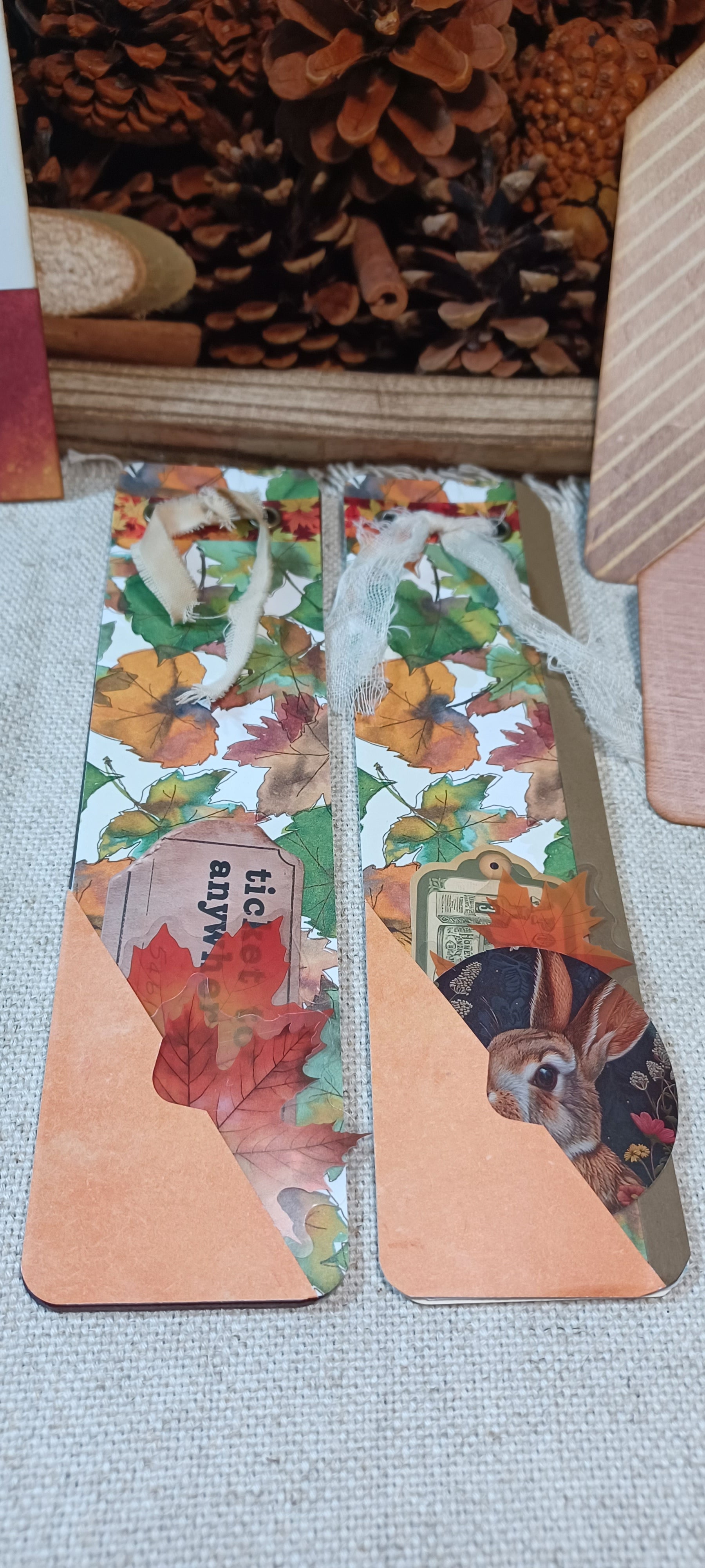 Two handmade paper bookmark holders with autumn leaf and animal collage patterns, featuring a peach-colored triangular bottom section and a white twine loop at the top, displayed on a textured gray fabric surface.
