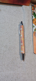 A clear plastic retractable pen with a floral pattern in orange, yellow, and purple, resting on a textured beige fabric surface.