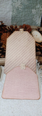A wooden decorative sign shaped like a house, featuring diagonal striped patterns on the upper section and a solid wood lower section, displayed on a textured fabric surface with rustic pinecones and a quilted background.
