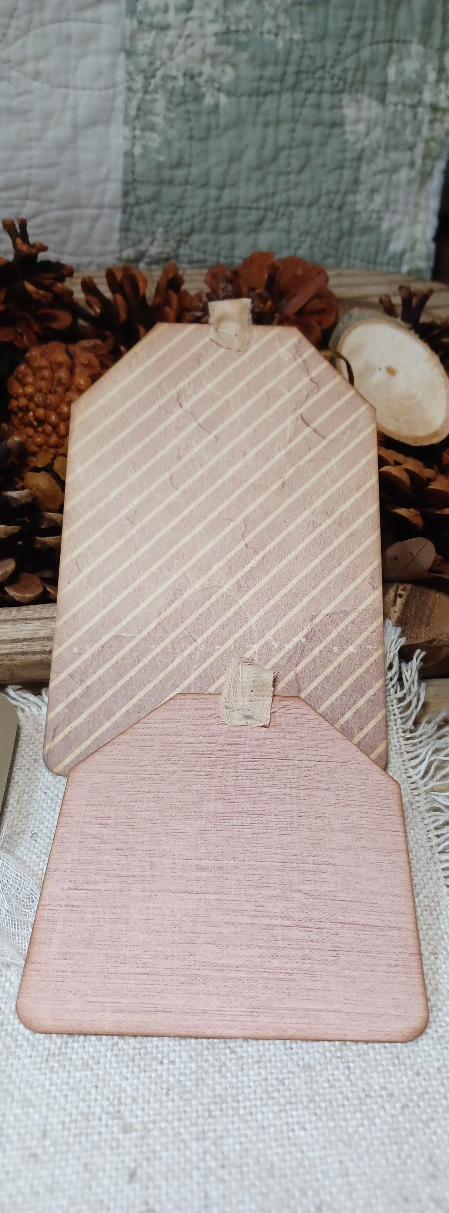 A wooden decorative sign shaped like a house, featuring diagonal striped patterns on the upper section and a solid wood lower section, displayed on a textured fabric surface with rustic pinecones and a quilted background.