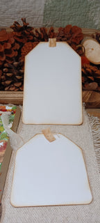 Two blank white gift tags with rustic brown borders and small wooden hangers, displayed on a burlap surface with pinecones and dried autumn foliage in the background.