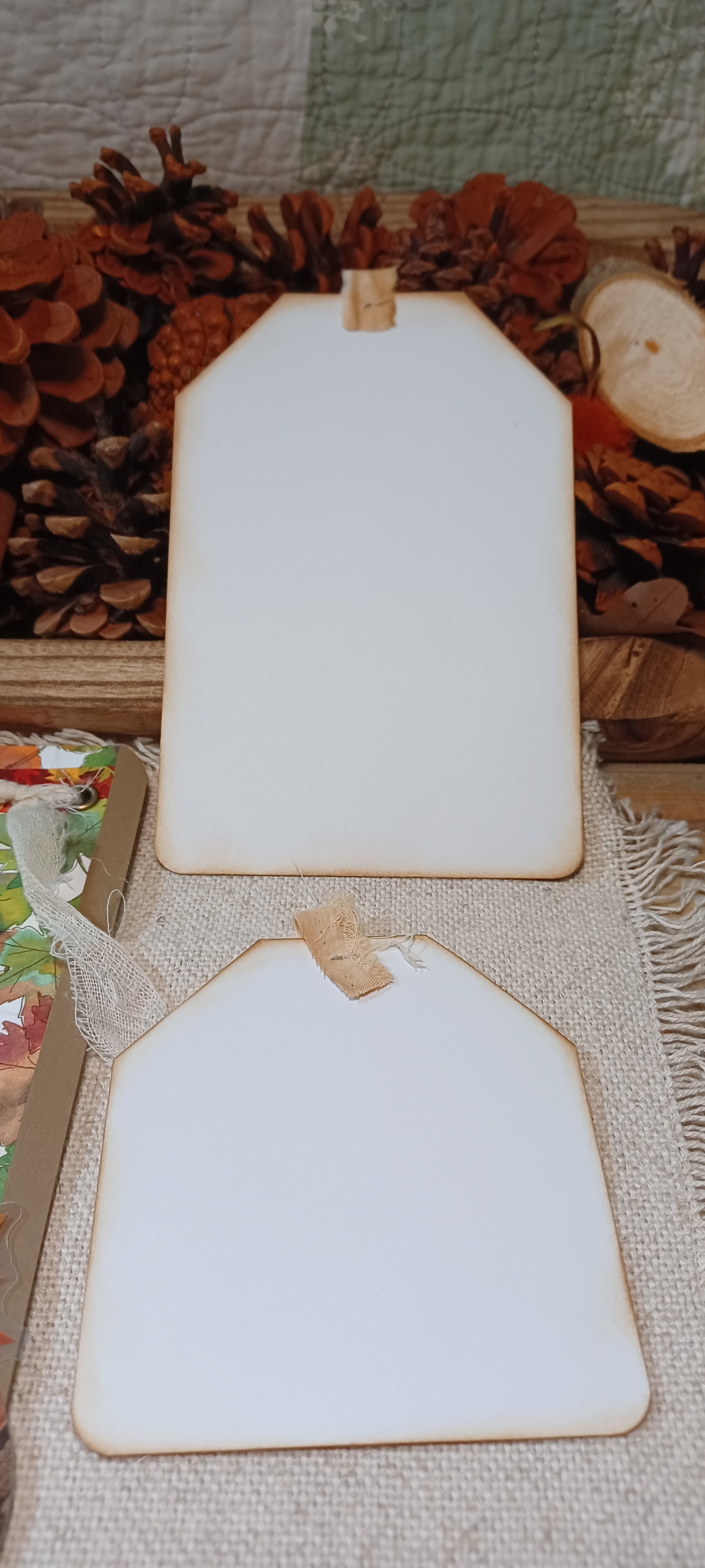 Two blank white gift tags with rustic brown borders and small wooden hangers, displayed on a burlap surface with pinecones and dried autumn foliage in the background.
