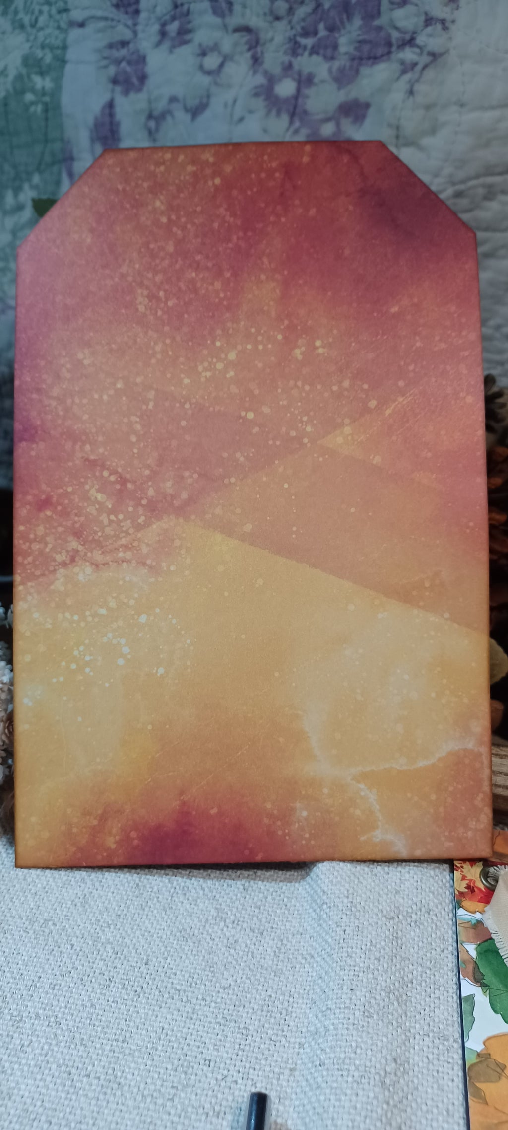 A rectangular art print or canvas with a stylized pointed top, featuring an abstract watercolor-like design in warm tones of orange, yellow, and reddish-brown, resting on a textured fabric surface.