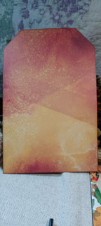 A rectangular art print or canvas with a stylized pointed top, featuring an abstract watercolor-like design in warm tones of orange, yellow, and reddish-brown, resting on a textured fabric surface.