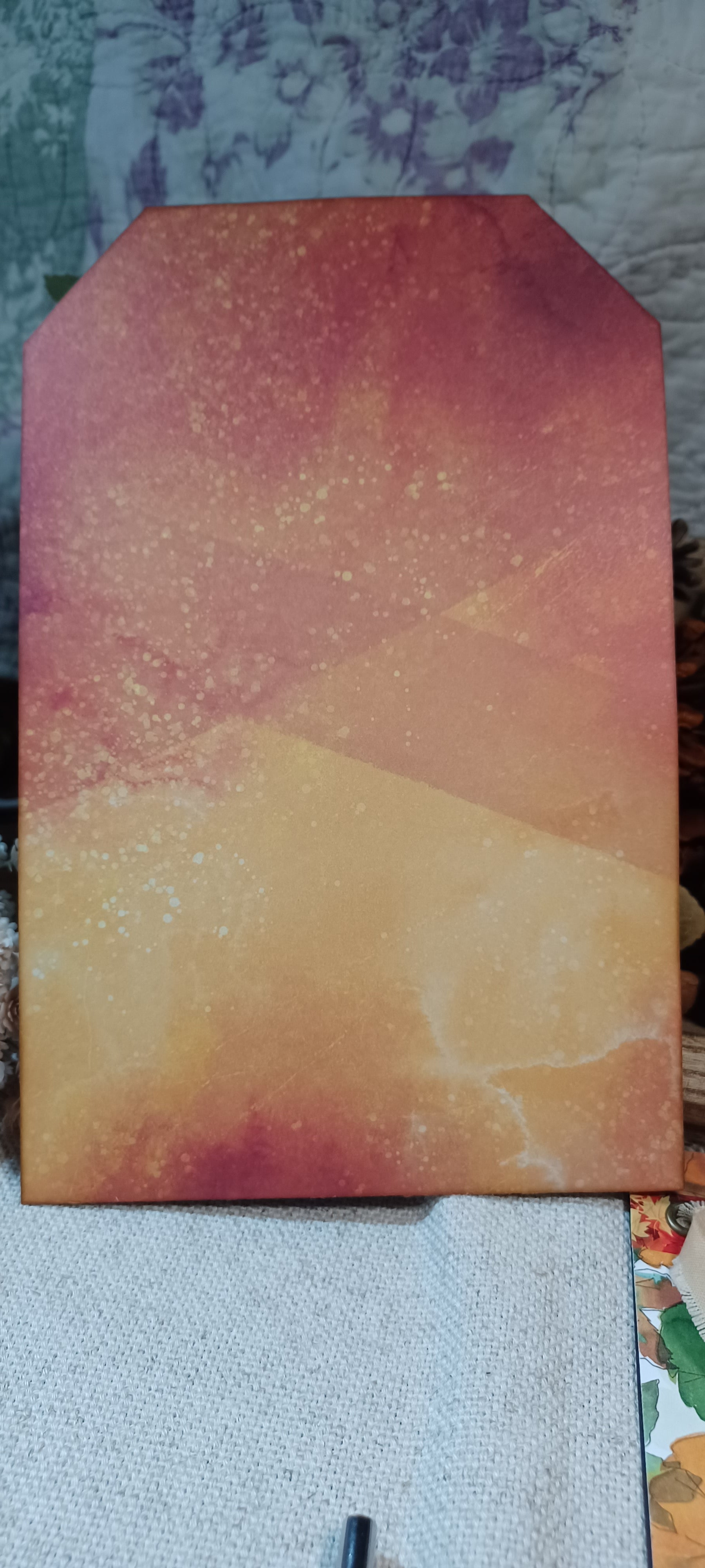A rectangular art print or canvas with a stylized pointed top, featuring an abstract watercolor-like design in warm tones of orange, yellow, and reddish-brown, resting on a textured fabric surface.