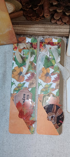 Two decorative paper bookmarks with an autumn leaf pattern and orange tabs, displayed on a textured fabric surface with pinecones in the background.