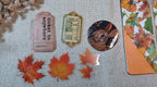 A flat-lay display of autumn-themed stationery and decorative items on a beige linen fabric background, including a pink vintage-style ticket, a green ornate label, a round pin with a rabbit illustration, and a notebook page decorated with colorful fall leaves.