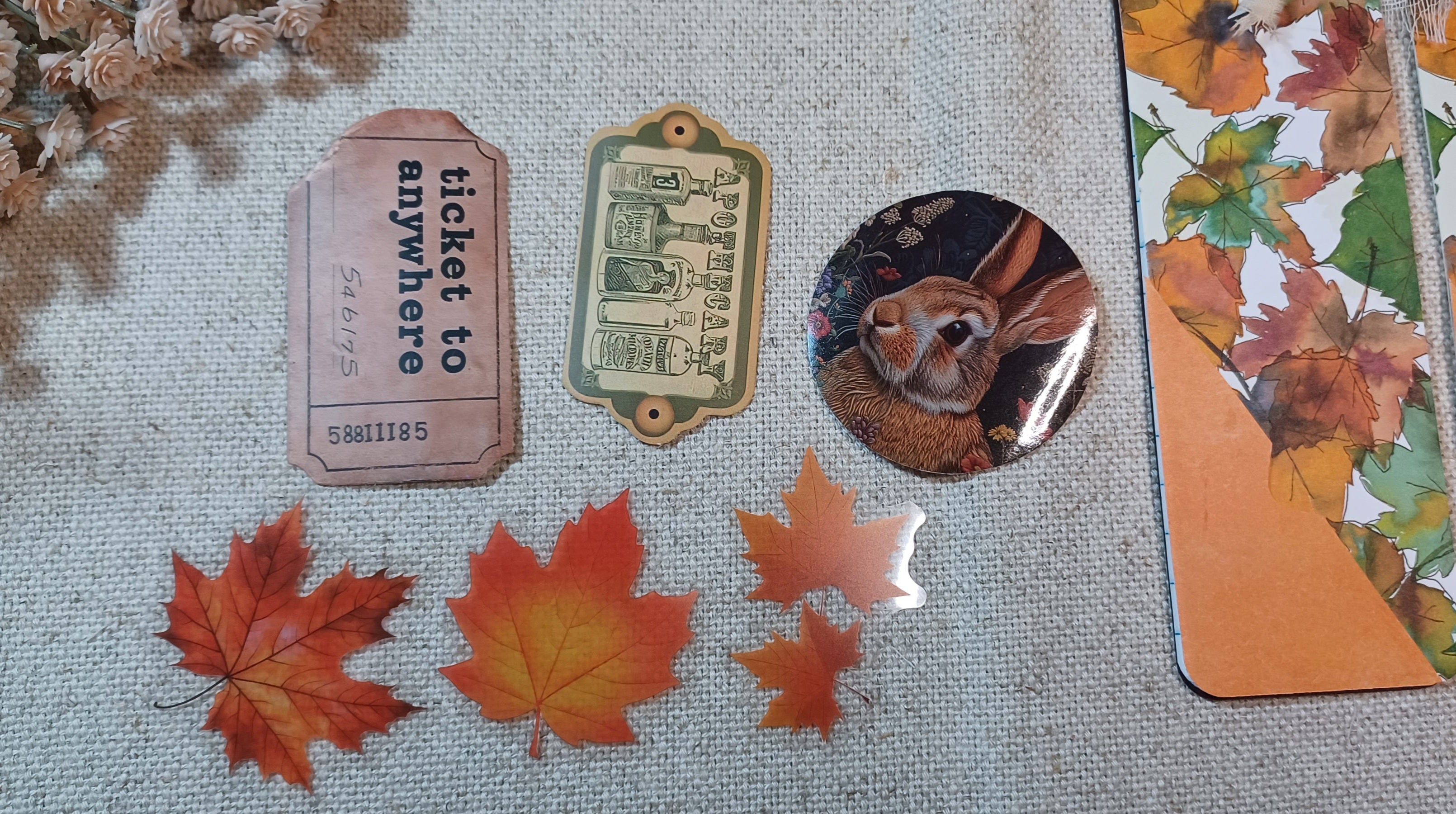 A flat-lay display of autumn-themed stationery and decorative items on a beige linen fabric background, including a pink vintage-style ticket, a green ornate label, a round pin with a rabbit illustration, and a notebook page decorated with colorful fall leaves.