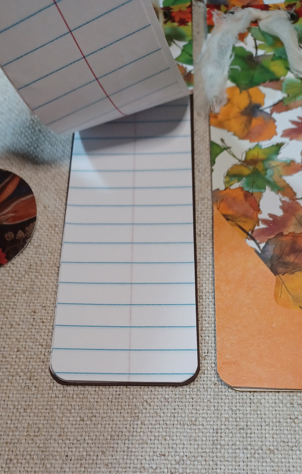 A close-up of a lined notepad page with blue and red lines, placed on a woven beige fabric surface next to a printed autumn leaf card or pad.