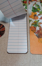 A close-up of a lined notepad page with blue and red lines, placed on a woven beige fabric surface next to a printed autumn leaf card or pad.