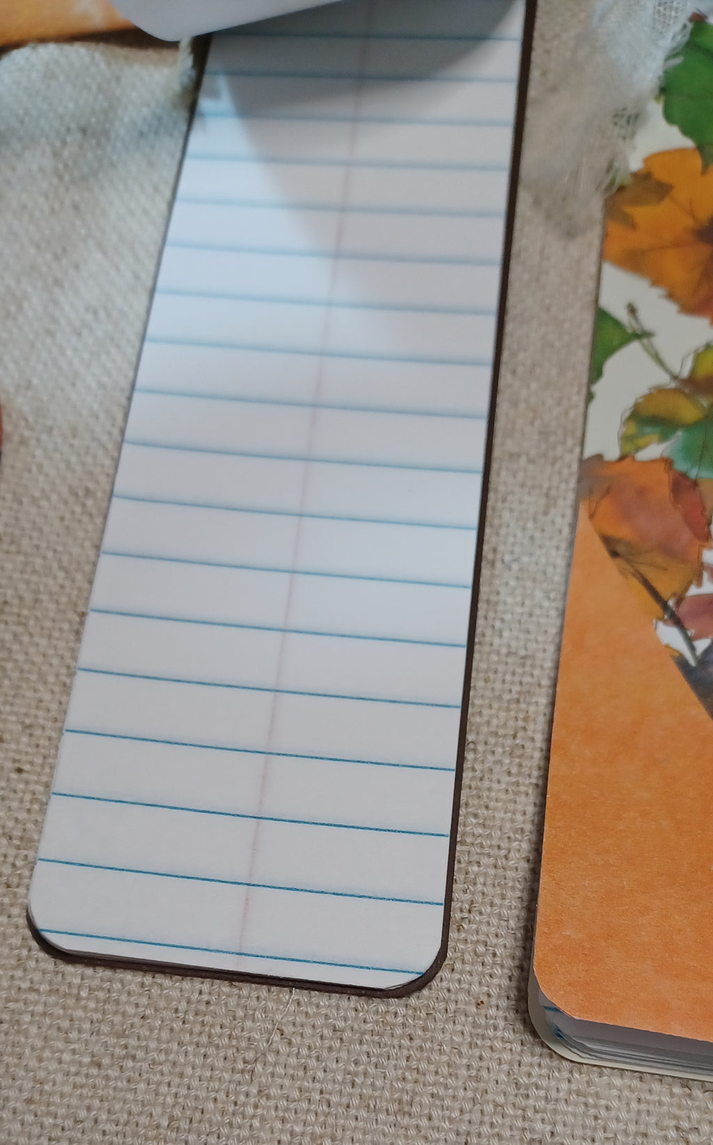 A close-up of a rectangular notepad or bookmark with blue horizontal lines on a white background, placed on a beige woven surface next to another orange-toned item.