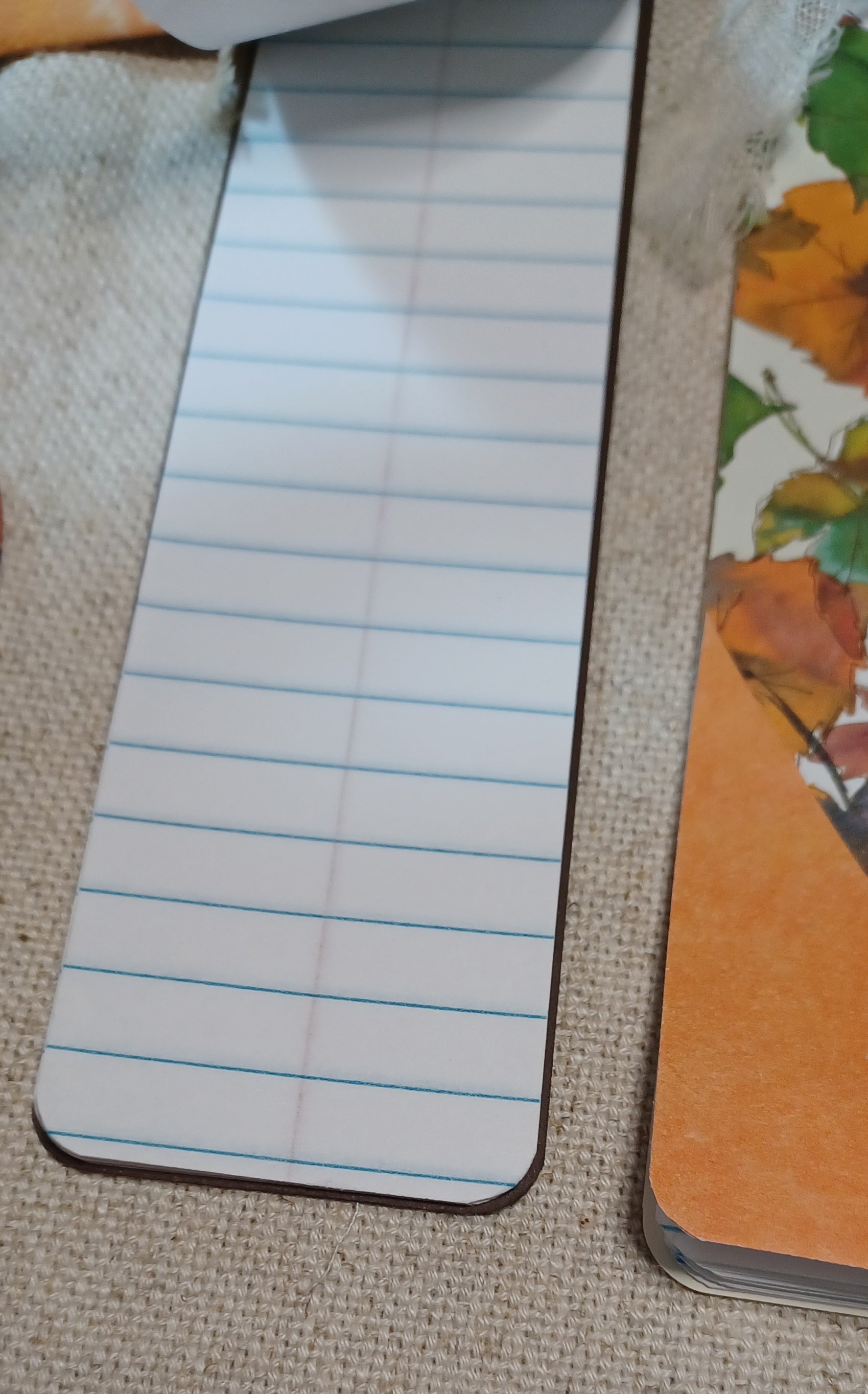 A close-up of a rectangular notepad or bookmark with blue horizontal lines on a white background, placed on a beige woven surface next to another orange-toned item.
