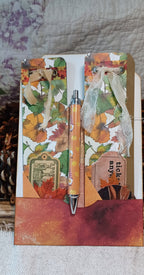 An autumn-themed handmade journal or notebook with a decorative pen, featuring colorful fall leaves, ribbons, and a textured orange-brown base, displayed on a neutral fabric background.