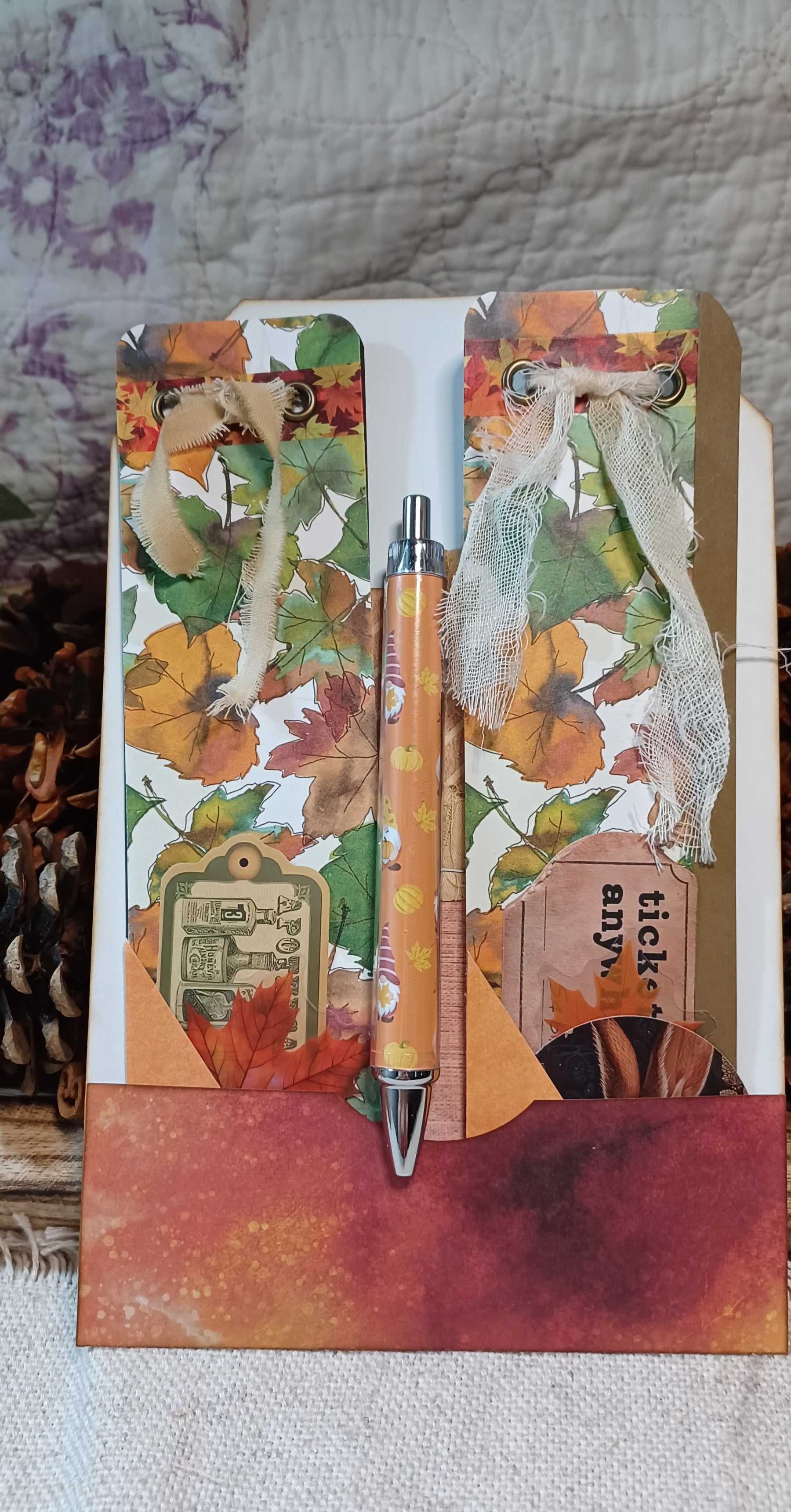 An autumn-themed handmade journal or notebook with a decorative pen, featuring colorful fall leaves, ribbons, and a textured orange-brown base, displayed on a neutral fabric background.