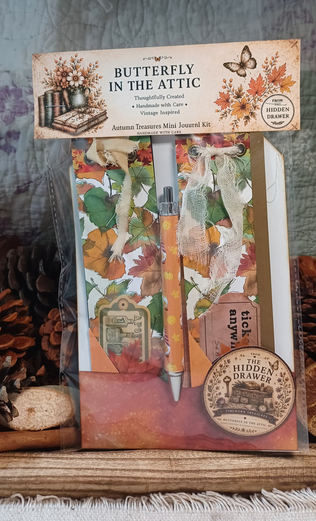 Butterfly in the Attic Autumn Treasure Mini Journal Kit in its clear plastic packaging, featuring vintage-style autumn illustrations, a pen, and a hidden drawer insert, displayed on a rustic wooden surface with pinecones.