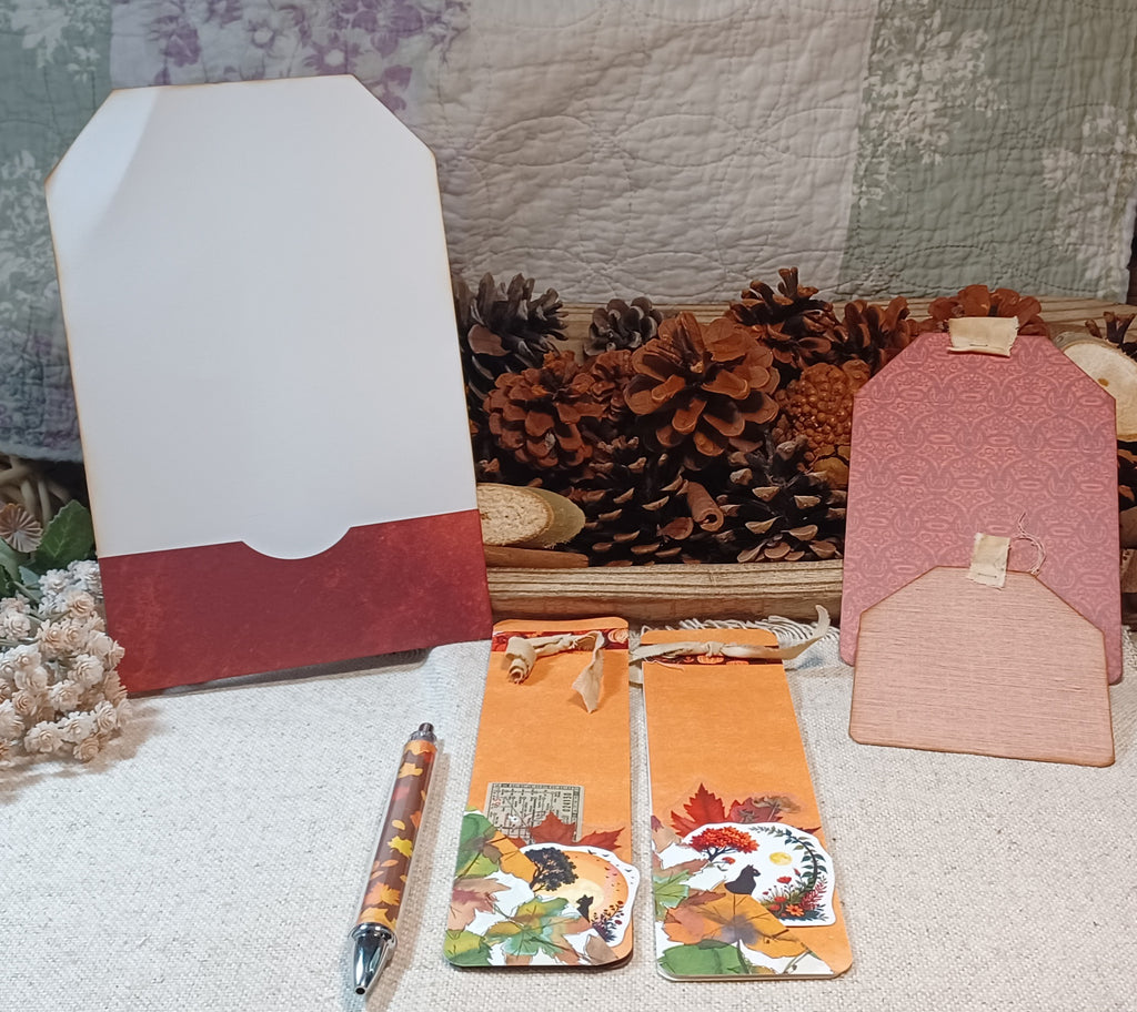 A styled flat-lay of autumn-themed stationery and decorative accessories on a textured cream fabric surface, featuring a white and red envelope, a pink patterned tag, a peach tag, a patterned pen, and two orange bookmarks with fall leaves and a dog illustration.