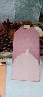 A pink decorative card or envelope-style paper piece with a patterned back and a plain front tab, displayed on a light fabric surface with pinecones and a quilted textile in the background.