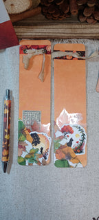 Two decorative paper bookmarks with orange-brown backgrounds, floral and botanical cutout designs, and ribbon ties, displayed on a textured fabric surface next to a patterned pen.