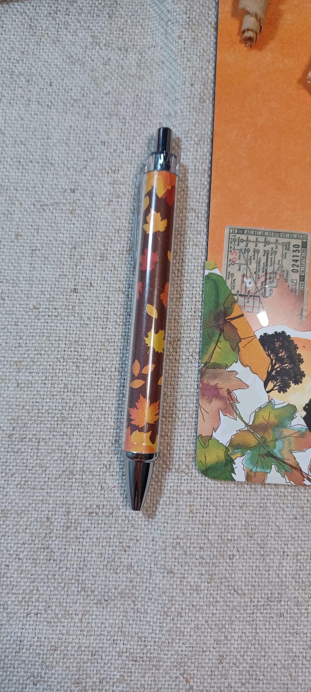 A slim ballpoint pen with a clear barrel showing an autumn leaf pattern in orange, yellow, and brown, with black accents, resting on a textured beige fabric surface next to an orange notebook.