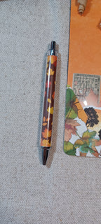 A slim ballpoint pen with a clear barrel showing an autumn leaf pattern in orange, yellow, and brown, with black accents, resting on a textured beige fabric surface next to an orange notebook.
