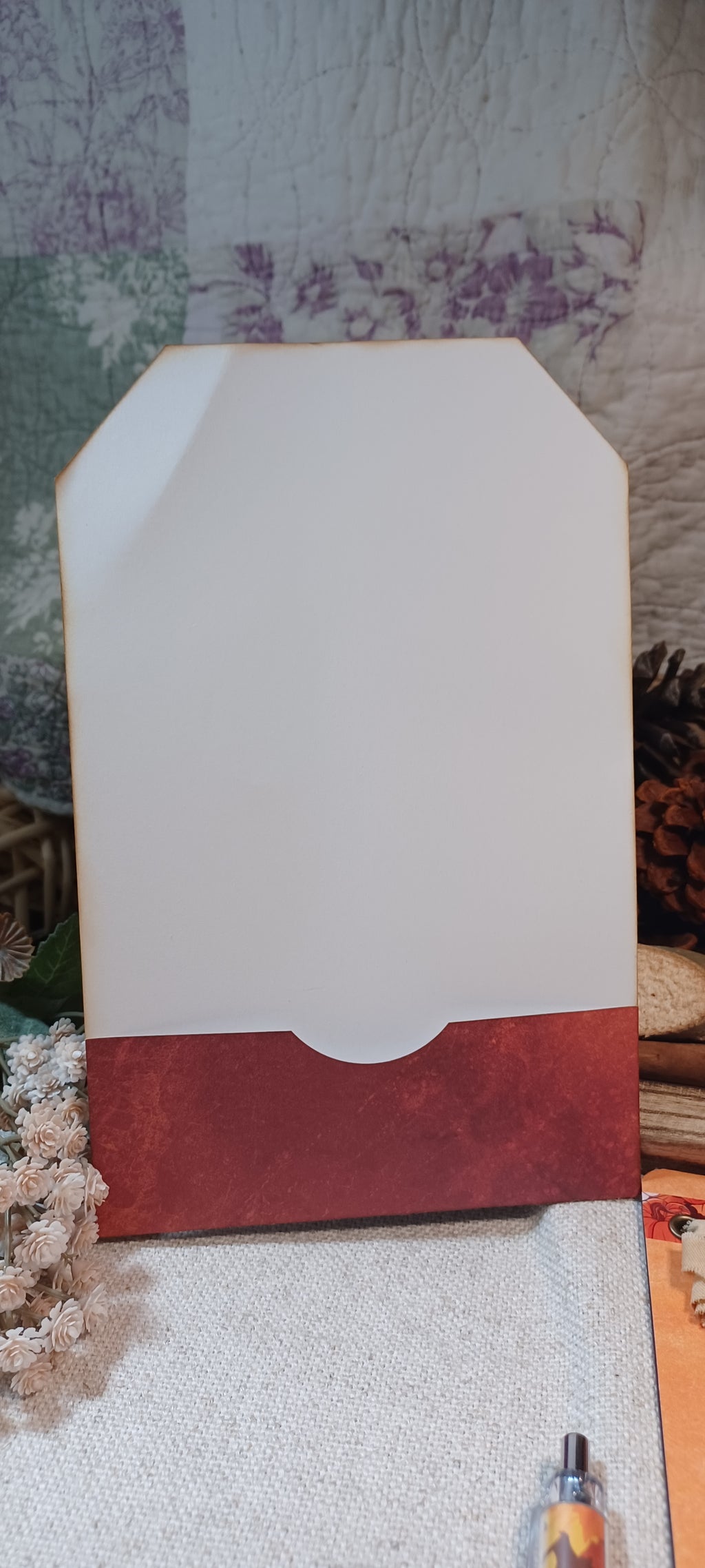 A handmade paper envelope or card holder with a white top flap and a textured rust-red bottom base, displayed on a light fabric surface with a rustic, patterned background.