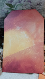 A rectangular art print or card featuring an abstract watercolor sunset design with a gradient of orange, yellow, and reddish-brown, displayed on a textured fabric surface.