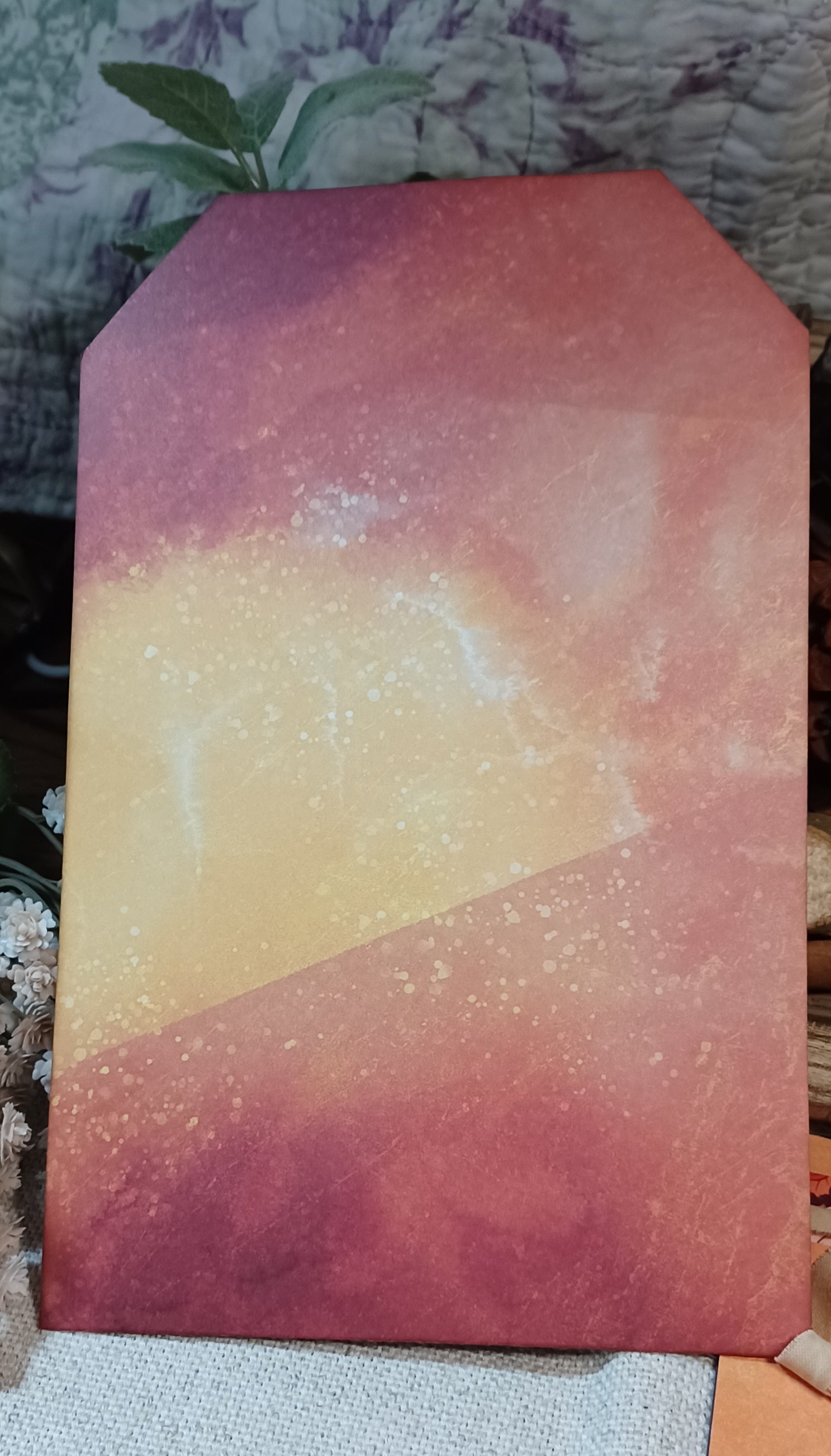 A rectangular art print or card featuring an abstract watercolor sunset design with a gradient of orange, yellow, and reddish-brown, displayed on a textured fabric surface.