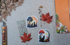 Two square die-cut stickers with autumn-themed cat silhouettes are displayed on a beige linen fabric alongside red maple leaves, dried flowers, a pen, and a notebook.