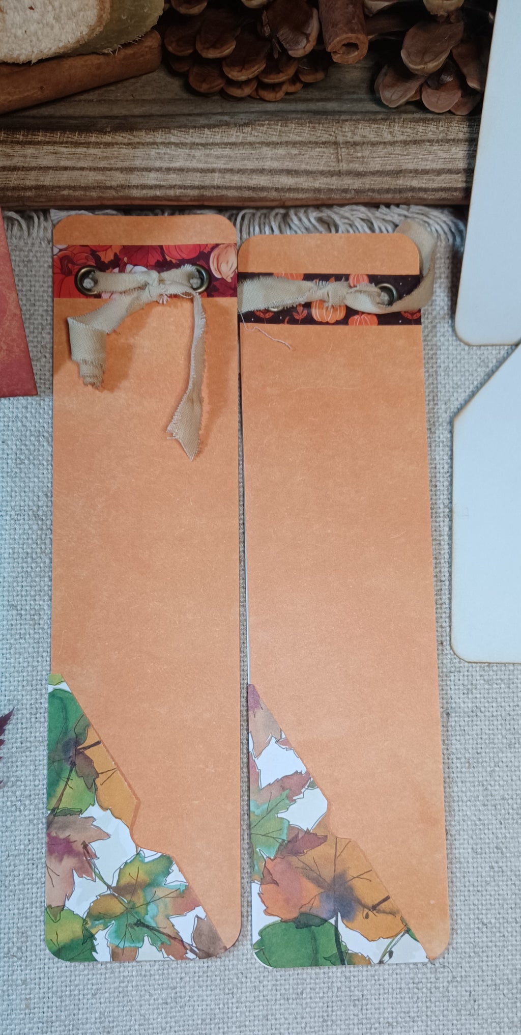 Two handmade paper bookmarks with autumn-themed designs, featuring a solid peach-orange center with printed fall leaves and pumpkins near the top and bottom, laid flat on a textured fabric surface.