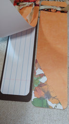 A close-up of a handmade bookmark or notebook page insert with a lined white paper section bordered in dark brown, an orange paper section, and a decorative leaf pattern along the bottom edge.