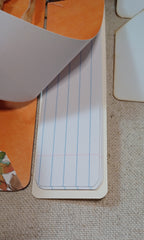 A close-up of a lined notepad page with blue vertical lines and a red margin line, partially lifted from an orange book or folder on a beige textured fabric surface.