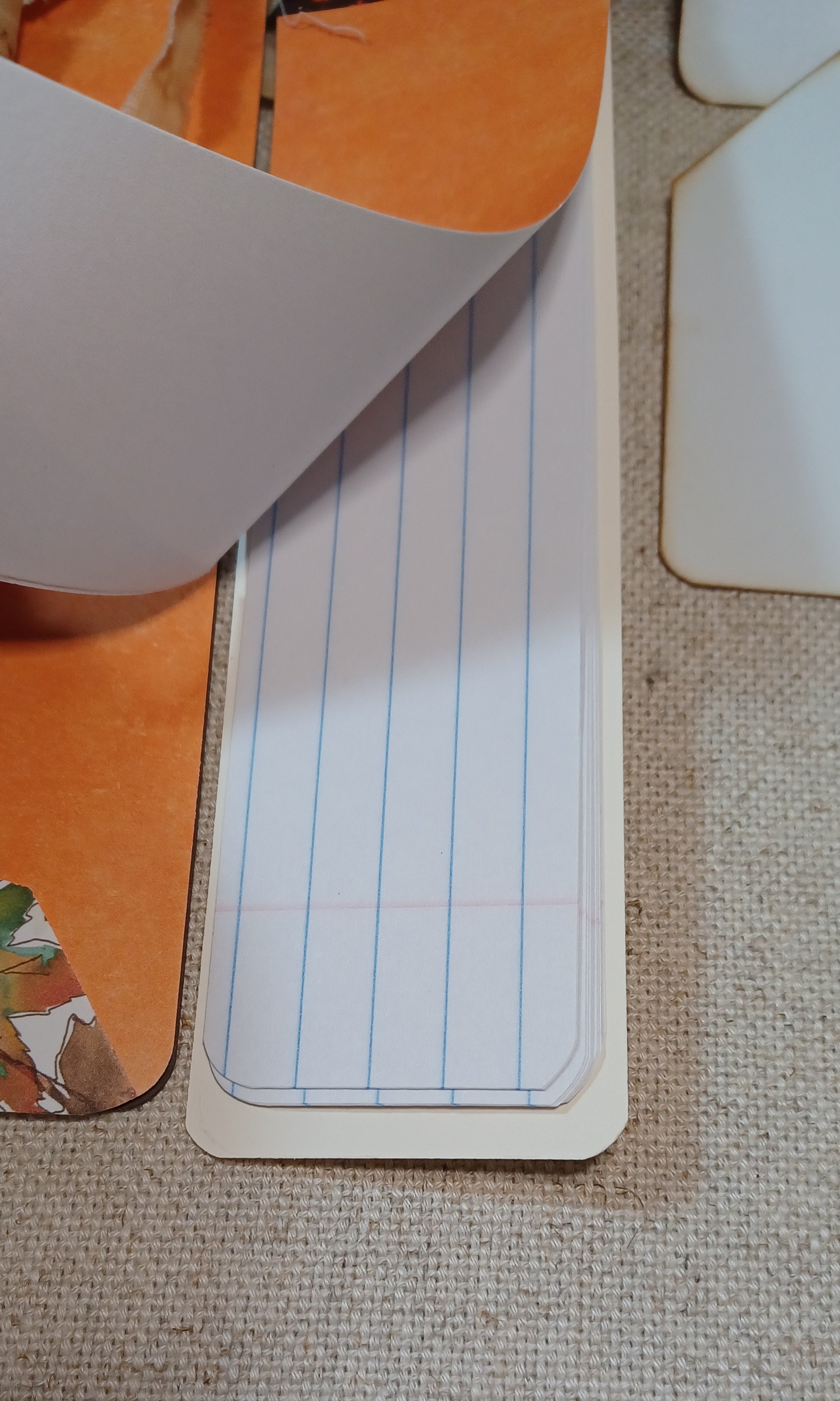 A close-up of a lined notepad page with blue vertical lines and a red margin line, partially lifted from an orange book or folder on a beige textured fabric surface.