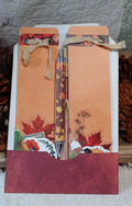 An autumn-themed pen gift set featuring a silver ballpoint pen with a fall leaf pattern, nestled inside a white envelope decorated with orange cardstock, red maple leaves, and a rustic red paper base.