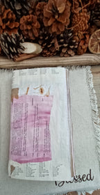 A handmade journal made from recycled paper, featuring a pink watercolor wash on the cover, resting on a fringed beige throw blanket with pinecones in the background.