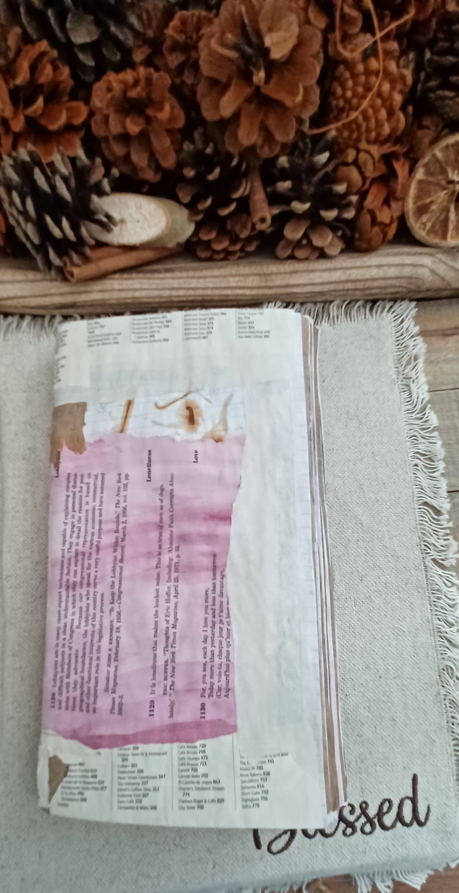 A handmade journal made from recycled paper, featuring a pink watercolor wash on the cover, resting on a fringed beige throw blanket with pinecones in the background.