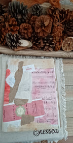A handmade fabric art piece featuring a collage of vintage pink sheet music, brown paper strips, and white fabric, finished with fringe and the word 'Blessed' stitched at the bottom. The piece is displayed on a wooden surface surrounded by pinecones and dried citrus slices.