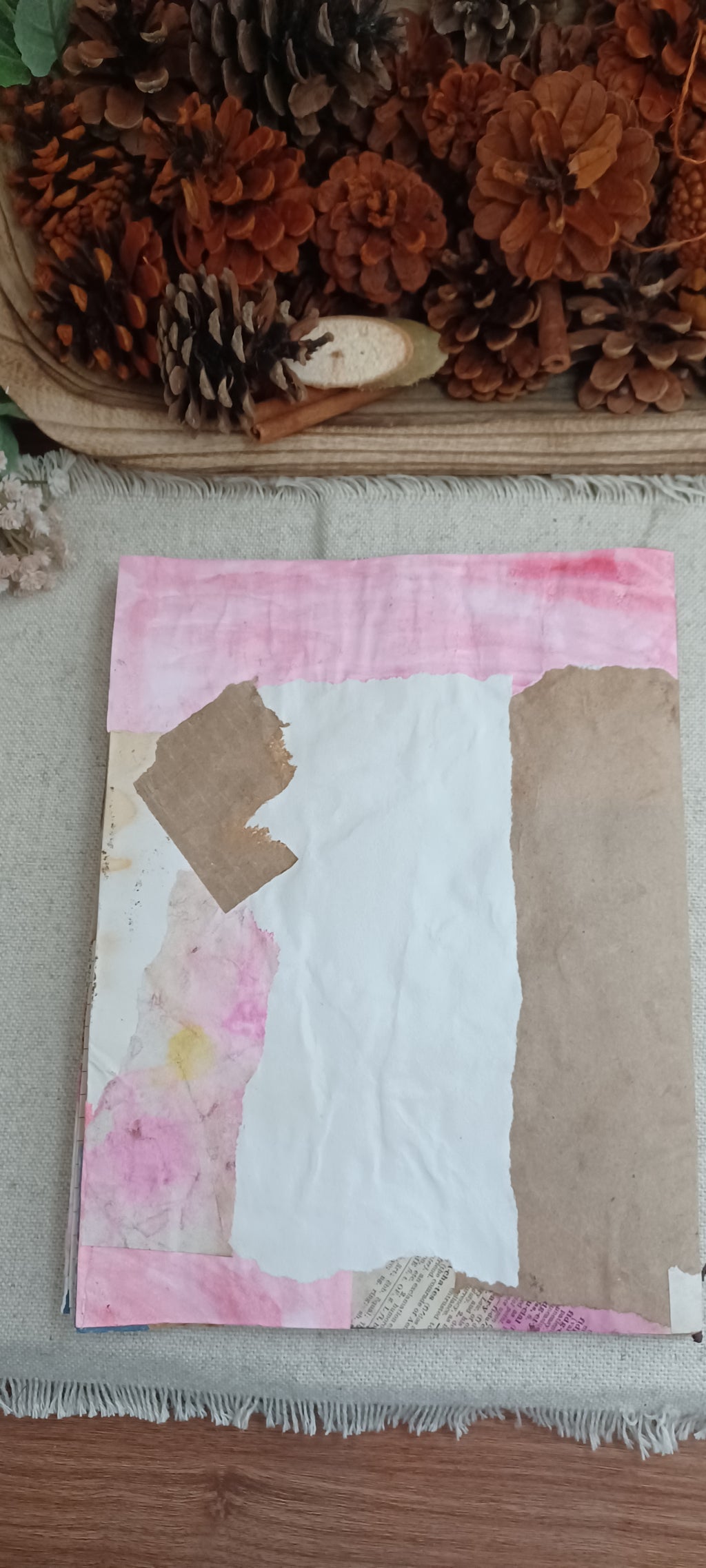 A handmade patchwork art piece featuring pink watercolor washes, white paper, and brown kraft paper sections, displayed on a textured fabric surface with pinecones in the background.