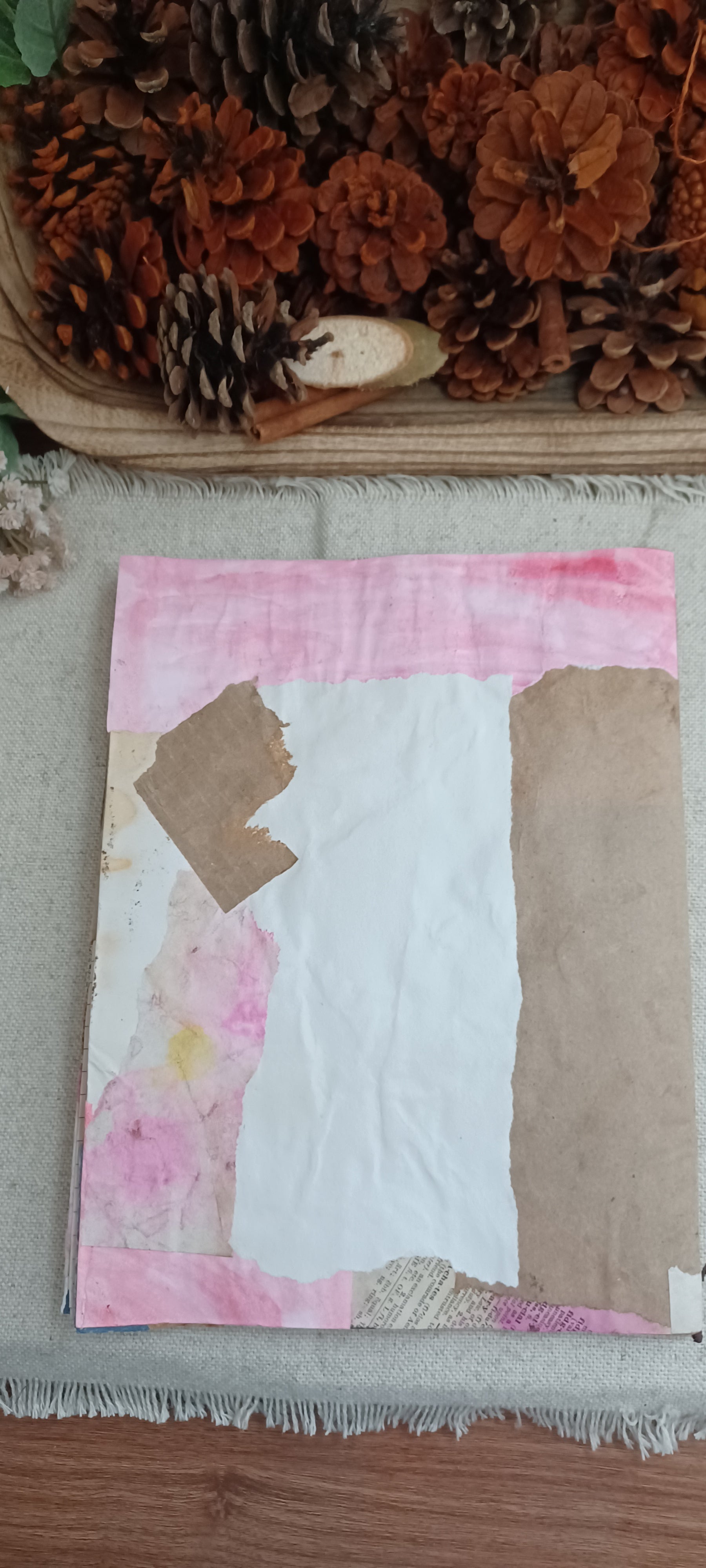 A handmade patchwork art piece featuring pink watercolor washes, white paper, and brown kraft paper sections, displayed on a textured fabric surface with pinecones in the background.