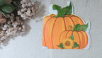 A   autumn napkin featuring two orange pumpkins with green leaves and a small yellow sunflower, placed on a light fabric surface with dried flowers at the side.