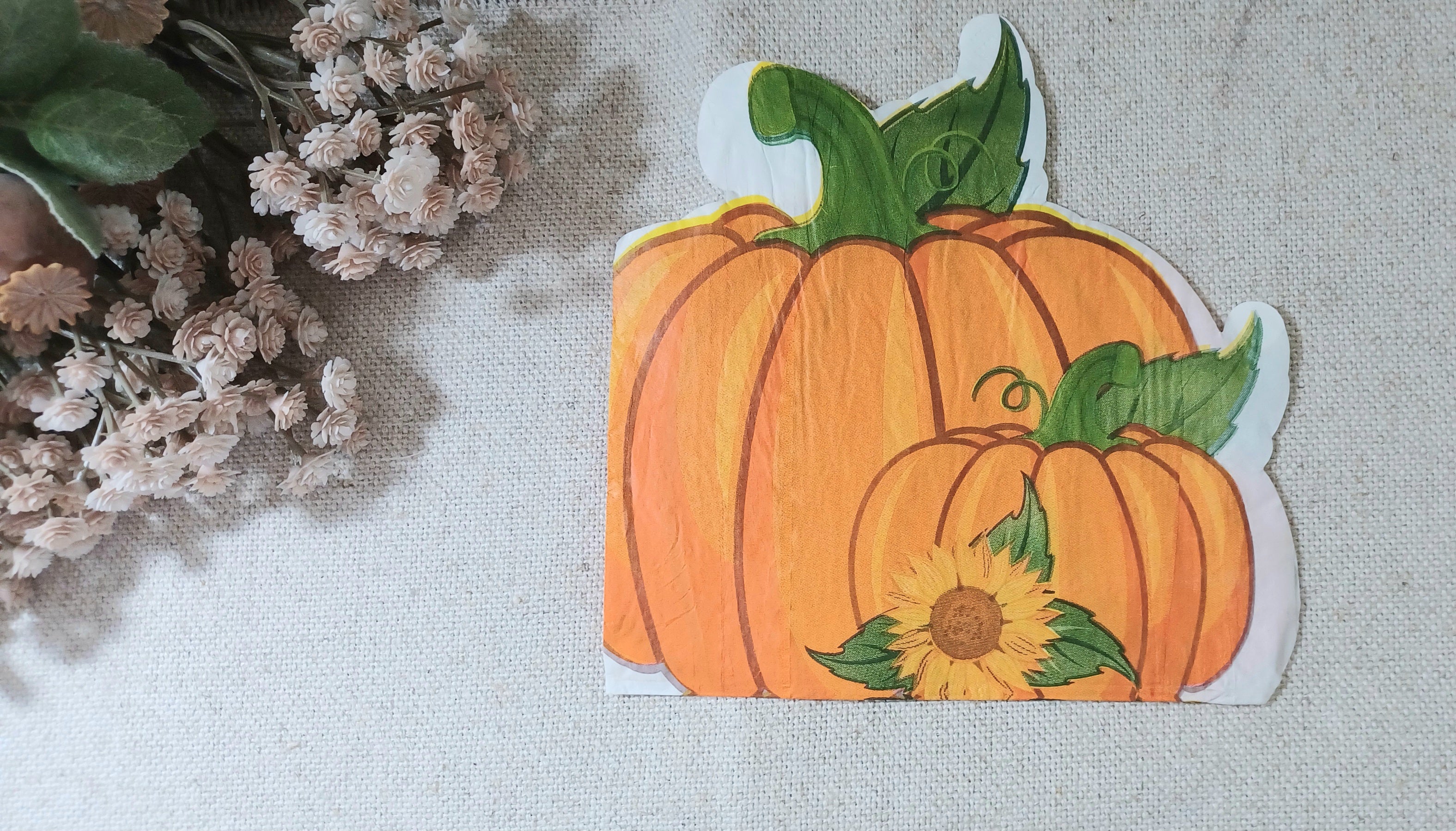 A   autumn napkin featuring two orange pumpkins with green leaves and a small yellow sunflower, placed on a light fabric surface with dried flowers at the side.
