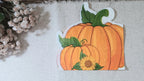 A die-cut paper  autumn napkin featuring two hand-painted orange pumpkins with green stems and a small sunflower, displayed on a textured beige fabric next to dried flowers.