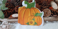 A  autumn napkin with two orange pumpkins with green stems and a small sunflower, displayed on a rustic wooden surface with pine cones and dried botanicals in the background.