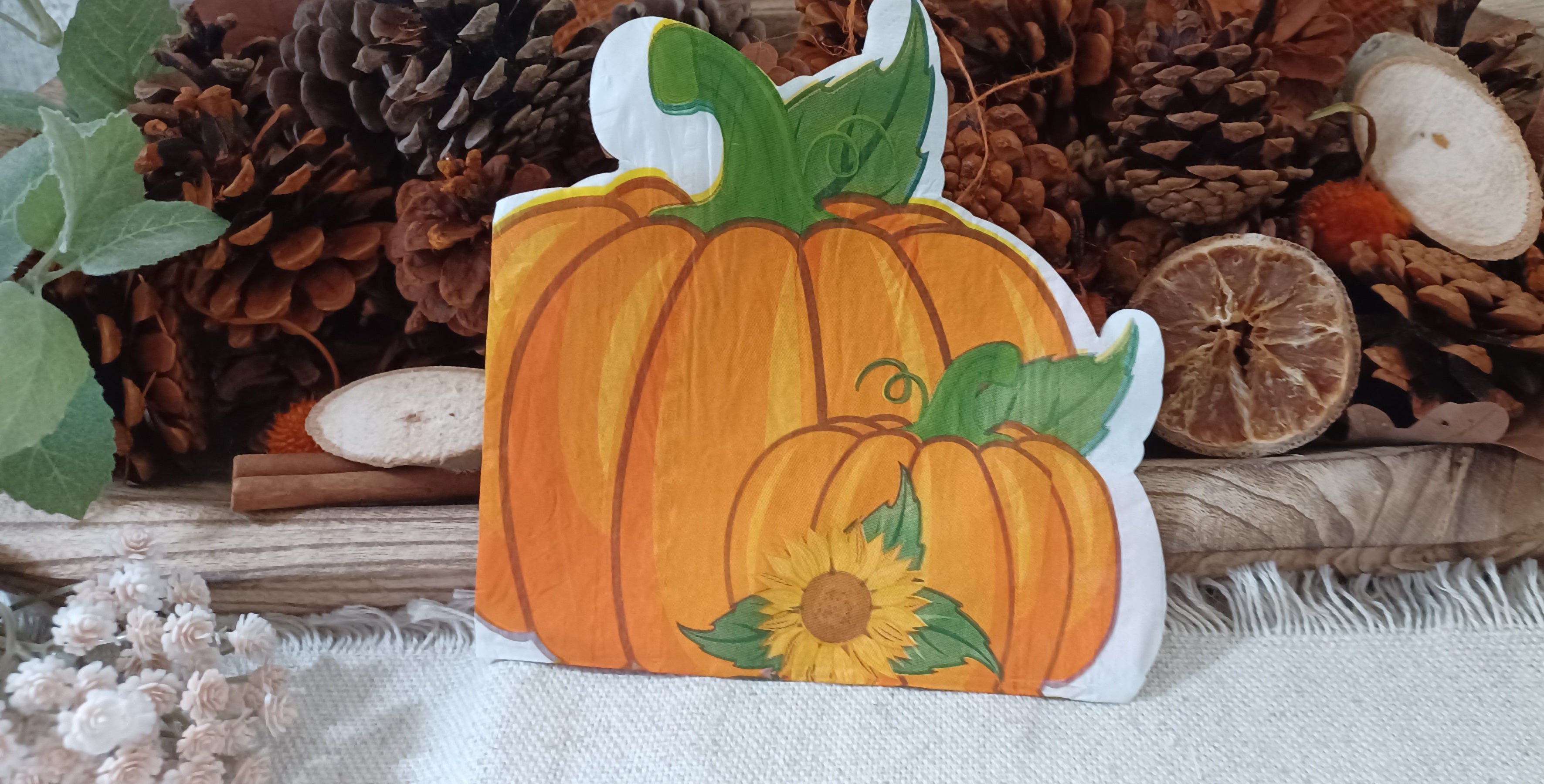 A  autumn napkin with two orange pumpkins with green stems and a small sunflower, displayed on a rustic wooden surface with pine cones and dried botanicals in the background.